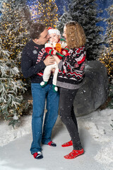 family Christmas moment with parents holding baby in snowy holiday setting. Both adults wear festive Christmas sweaters, while baby is dressed in a cute holiday outfit with Santa hat. Snowflakes fall 