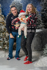 Happy family Christmas portrait in snowy winter setting. Mother and father wearing festive holiday sweaters stand together holding baby dressed in a Santa hat and Christmas outfit