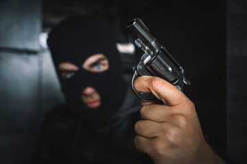 Terrorist hooded in balaclava, holding gun in hand. Man aiming with a pistol, dark building indoor background, Killer, armed robbery concept.