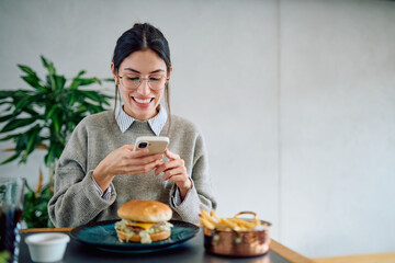 Woman smiling while taking a picture of her burger and fries with a smartphone at a restaurant,...
