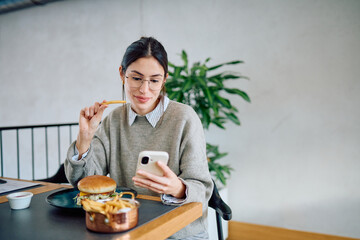 Young woman enjoying a fast-food meal while using her mobile phone, multitasking during a casual lunch