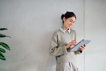 Smiling young woman wearing casual business attire and glasses, reading or typing on a digital tablet in a modern office
