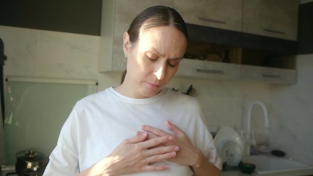 Young woman feeling unwell and suffering from heart pain, taking medication pill for treatment while standing in her kitchen, showing symptoms of cardiovascular disease or panic attack. Heart attack