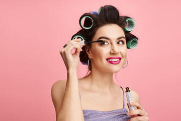 Young woman applying mascara with curlers in her hair against a vibrant pink background