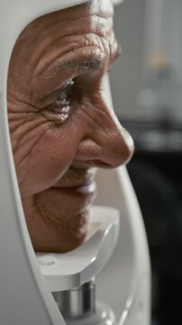 Vertical closeup shot of grey haired elderly woman smiling softly while putting head into frame of modern diagnostic machine during eye exam