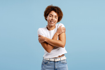 Young woman embracing self, feeling content and practicing self care on blue background
