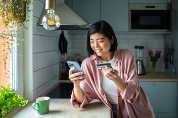 Adult woman smiling, paying by credit card on smartphone in kitchen