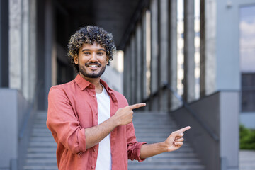 Portrait of a smiling young Indian man standing outside and pointing with his fingers at a brown spot to the side