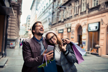 Young adult couple laughing at smartphone while shopping in city
