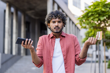 Close-up portrait of a young worried Indian man standing outside having problems with his credit card and phone