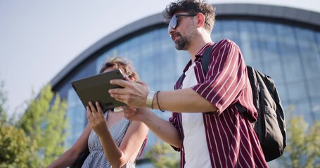 Smiling young couple using a tablet outdoors on a sunny day in an urban setting with modern architecture in the background