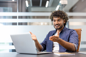 Obraz premium Smiling young Indian man sitting at a desk and chatting on a video call, holding an online meeting