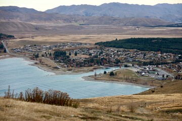 Obraz premium Mt John Observatory, Lake Tekapo, NZ - May 25, 2025: High-angle panoramic view of Lake Tekapo township nestled along turquoise water. The town is surrounded by the wide, brown hills of Mackenzie Basin