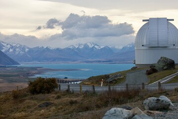 Mt John Observatory, Lake Tekapo, NZ - May 25, 2025: A telescope dome sits on hill overlooking vast landscape. Turquoise lake, snow-capped mountains of the Mackenzie Basin under a cloudy sky.