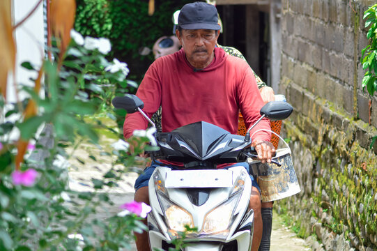 A front close-up view of a middle-aged villager couple riding their motorbike into a narrow residential path, the father drives forward while the mother rides pillion carrying a bucket.