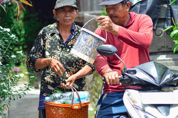 A middle-aged villager couple stands in their yard after arriving on their motorbike, the father holds a dirty bucket while the mother carries an orange basket with a bottle and leaves.