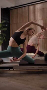 Engaging Pilates class shows two women performing a series of exercises on reformers in a spacious studio with natural light. Focus on flexibility and strength