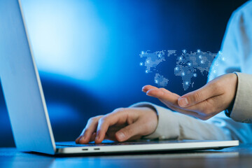 Hands interact with a glowing digital world map above a laptop at a desk in a modern work environment