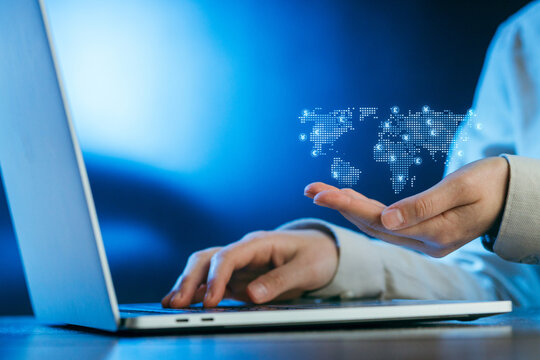 Hands interact with a glowing digital world map above a laptop at a desk in a modern work environment