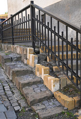 Stairs showing signs of wear with uneven bricks and a black railing in an urban setting