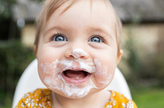 portrait of face, happy little girl under one year old, eating yogurt on her own and getting dirty. child's face is smeared with food. Happy, sincere joy, smile. A cheerful childhood. Daily routine.