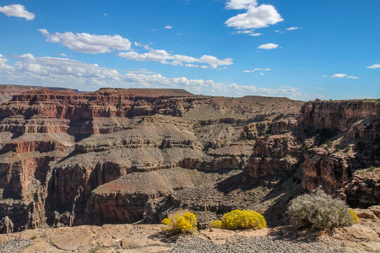 A panoramic view of the Grand Canyon's desert landscape, featuring layered rock formations, deep cliffs, and a vibrant blue sky.