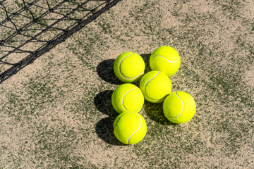 Six tennis balls grouped on sandy court surface beside black net; warm light and soft focus frame padel, tennis training, sportlife lifestyle and recreational play.