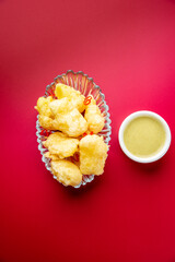 Crispy golden deep-fried chicken nuggets (or tempura pieces) served in a glass dish with a side of yellow dipping sauce on a vibrant red background, shot from above.