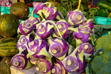 A bright close-up of a pile of fresh, purple kohlrabi bulbs displayed for sale on a market stall next to melons and broccoli.