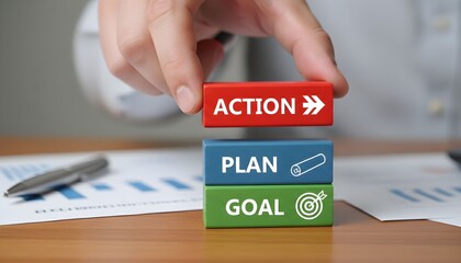A hand stacking wooden blocks labeled "ACTION", "PLAN", and "GOAL" on a desk with charts.