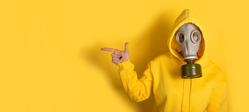 Individual in yellow hoodie and gas mask points finger on vibrant yellow background, with copy space for creative use.
