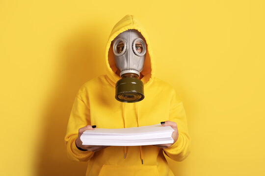 Individual in yellow hoodie and gas mask holding a stack of papers against a yellow background, emphasizing caution and information.