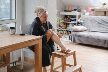 Woman applying coconut oil to leg in modern bedroom, natural beauty treatment and skincare routine...