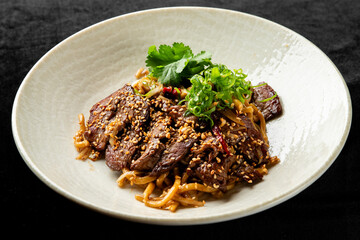 Gourmet Asian beef noodle stir-fry dish with sliced steak, thick noodles, sesame seeds, fresh cilantro, and green onions in a white bowl on a black background