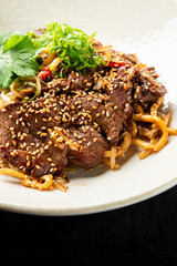 Gourmet Asian beef noodle stir-fry dish with sliced steak, thick noodles, sesame seeds, fresh cilantro, and green onions in a white bowl on a black background