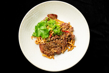 Gourmet Asian beef noodle stir-fry dish with sliced steak, thick noodles, sesame seeds, fresh cilantro, and green onions in a white bowl on a black background