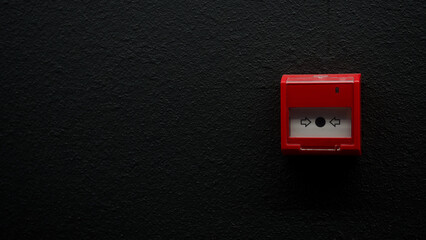Red manual fire alarm call point mounted on a modern textured black wall. Emergency safety button behind glass with negative space for text. Photo