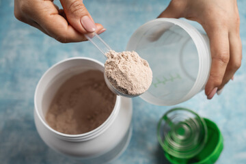 Close-up of female hand putting scoop of chocolate whey protein powder into shaker on blue background with plastic protein jar, concept of sport nutrition