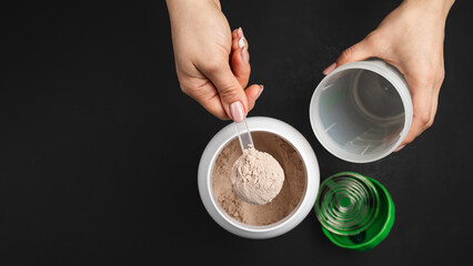 Top view of female hand putting scoop of chocolate whey protein powder into shaker with plastic...