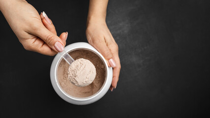 Fototapeta premium Top view of female hand holding measuring scoop with chocolate whey protein powder above plastic jar on dark background with copy space, concept of sports nutrition and dietary supplements