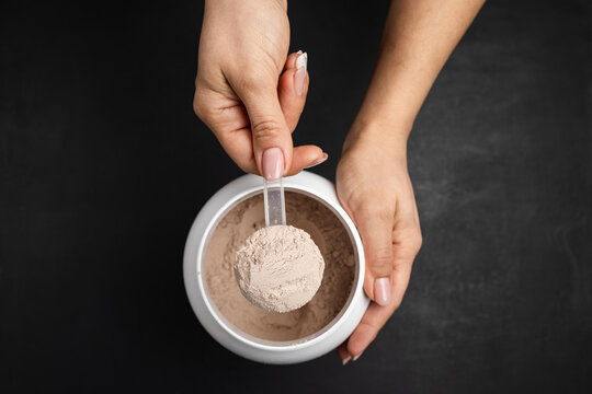 Top view of female hand holding measuring scoop with chocolate whey protein powder above plastic jar on dark background, concept of sports nutrition and dietary supplements