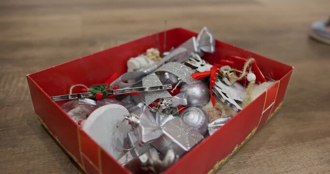 Woman hand unpacking red Christmas decoration box with silver ornaments, ribbons, baubles and festive toys. preparing holiday decorations, cozy seasonal home setup. Christmas tree decorations