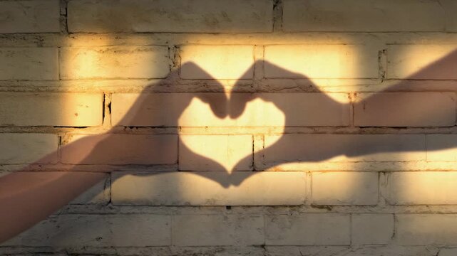 Warm golden sunlight casts a beautiful hand heart shadow against a textured rustic white brick wall backdrop