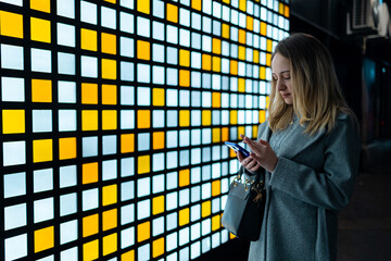 Young woman uses smartphone while standing next to a colorful wall at night in the city