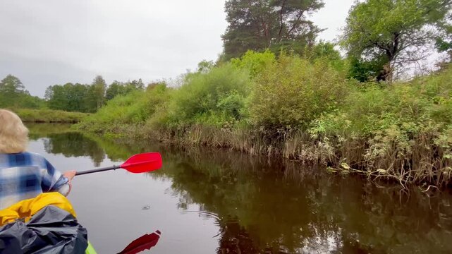 Slow canoe stroke moves the boat along Irbe River in Latvia, gliding beside dense riverside vegetation and calm dark water that reflects the overcast sky