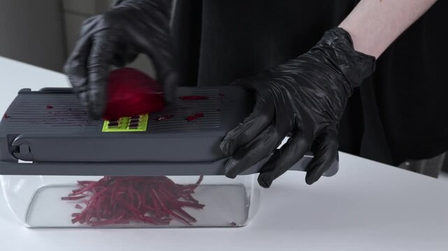 A person wearing black protective gloves skillfully shreds a whole fresh beetroot with a modern mandoline slicer, promoting safety and efficiency in healthy food preparation