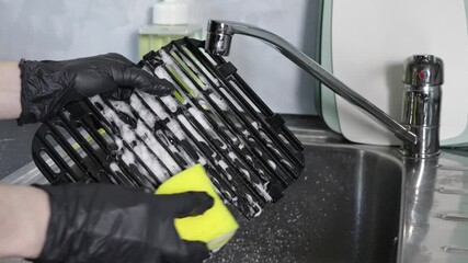 A person wearing black gloves vigorously scrubs a greasy air fryer grill grate with a bright yellow sponge, ensuring kitchen equipment cleanliness and essential domestic hygiene practices