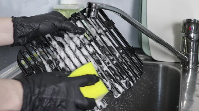 Hands in black gloves diligently wash a greasy air fryer rack using a yellow cleaning sponge and sudsy soap in a kitchen sink, ensuring cooking appliance hygiene