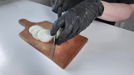 Person wearing black gloves precisely slices fresh white onion on a rustic wooden board, emphasizing hygienic food preparation and healthy cooking practices