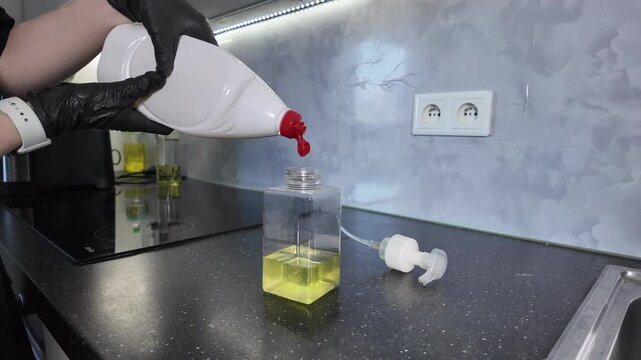 Person in black glove refilling a clear square dispenser bottle with yellow liquid soap on a modern kitchen counter, promoting efficient home cleaning and hygiene